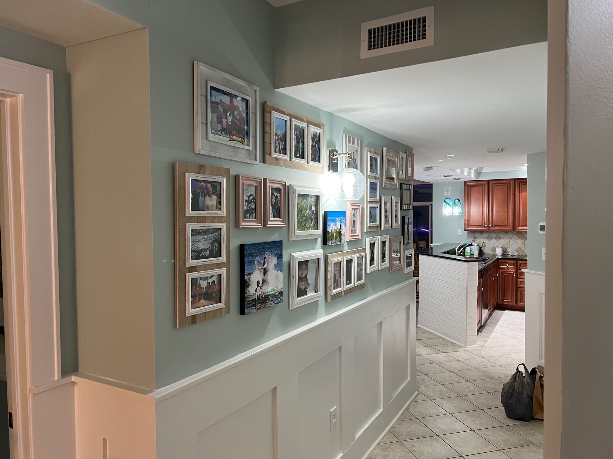 Un pasillo con una pared decorada con cuadros enmarcados, una zona de cocina con armarios de madera y un suelo de baldosas.