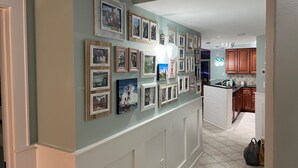 A hallway with a wall adorned with framed pictures, a kitchen area with wooden cabinets, and a tiled floor. - Luxury Penthouse 4BR Condo - Great for Families! (North Myrtle Beach)