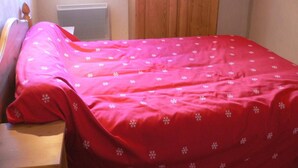 Quarto com uma cama coberta por um cobertor vermelho com desenhos de flocos de neve, cabeceira de madeira, radiador e porta de madeira.