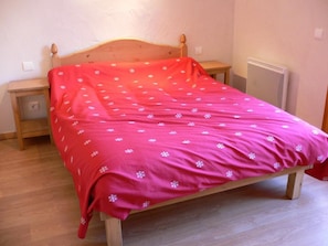 A single bed with a wooden frame and a red bedspread with white snowflake patterns. There is a wooden nightstand to the left of the bed with a small drawer. A radiator is visible to the right of the bed.