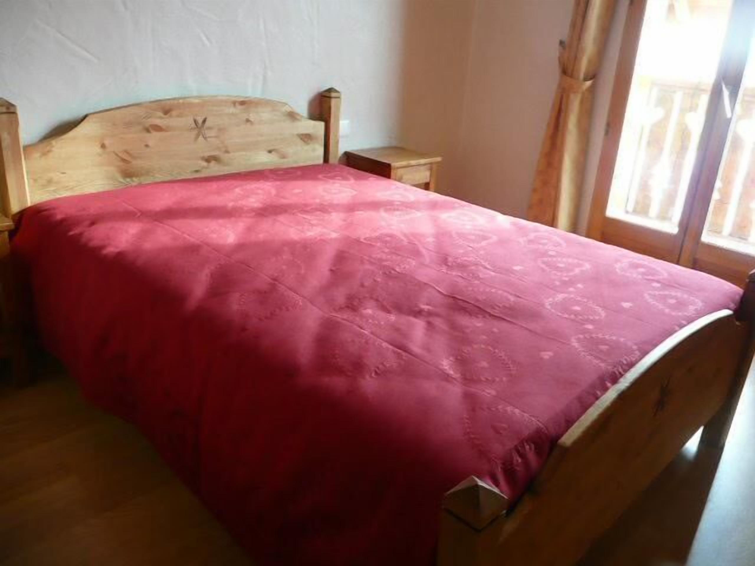 Un lit simple avec une tête de lit en bois et une couverture rouge, dans une pièce dotée d’un plancher en bois et d’une fenêtre avec des rideaux.