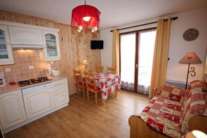 Een gezellige keuken met houten kastjes, een eettafel met stoelen, een rode hanglamp en uitzicht op de omgeving door een schuifdeur.