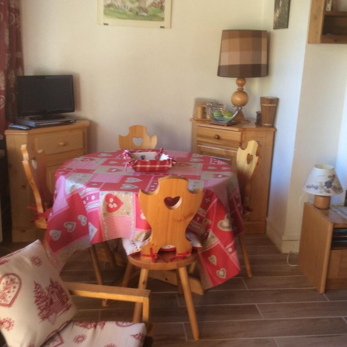 A dining area with a table covered in a red and white checkered tablecloth, wooden chairs, a wooden side table with a lamp, and a television on a wooden cabinet.
