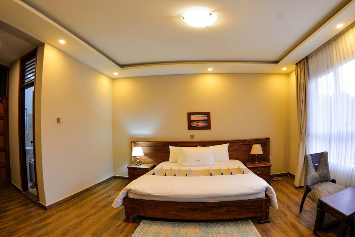 Une chambre d’hôtel avec un grand lit, deux tables de chevet, une chaise, une fenêtre avec des rideaux et un luminaire au plafond.