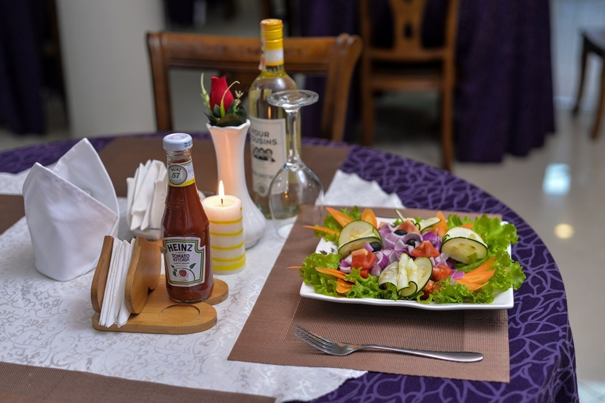 A table set with a salad, a bottle of Heinz tomato ketchup, a candle, and a bottle of olive oil.