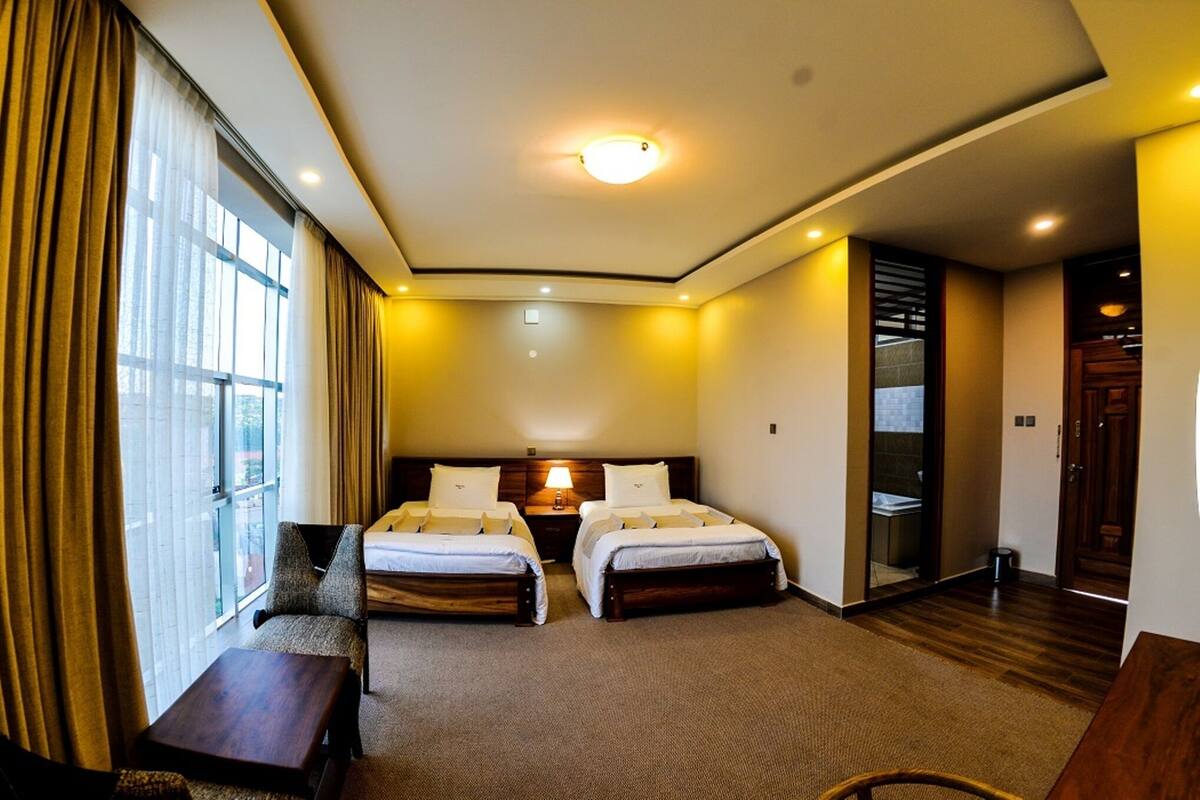 Une chambre d’hôtel avec deux lits, un bureau et une chaise. Il y a une fenêtre avec des rideaux, un luminaire au plafond et une porte qui donne sur une autre pièce.