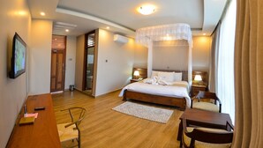A hotel room with a bed, bedside tables, a desk, a chair, a patterned rug, and a window with curtains. - Maple Leaf Hotel (Masaka)