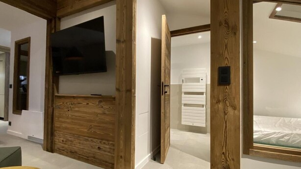 Ein modernes Hotelzimmer mit einer Wand in Holzoptik, einem Flachbildfernseher, einem Bett mit weißem Kopfteil und einer kleinen Sitzecke.