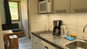 Et kompakt køkken med mikroovn, kaffemaskine og vask. En træbænk og et lille bord er synlige i det tilstødende opholdsområde.