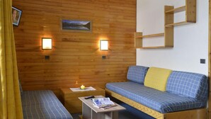 Habitación pequeña con pared de madera, dos camas con ropa de cama azul, una mesita con una vela y dos luces fijadas en la pared.