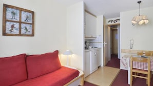 Une pièce de taille modeste, avec un canapé rouge, une petite table à manger recouverte d’une nappe à carreaux, un coin cuisine doté de meubles blancs et un téléphone fixé au mur.