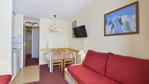 Une pièce de taille modeste, avec un canapé rouge, une table à manger en bois recouverte d’une nappe à carreaux, un petit coin cuisine et un téléviseur fixé au mur.