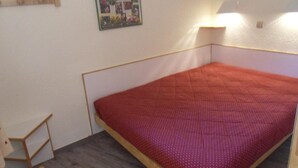 A small, single-bed room with a red bedspread, a wooden headboard, a wall-mounted mirror, and a framed collage of photos. - 100m from resort center, 6th floor, view ski slopes, balcony, tv, 40m², La Plagne (La Plagne)
