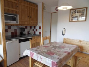 A compact kitchen with wooden cabinets, a microwave, and a dishwasher. There is a dining table with a checkered tablecloth and a wooden headboard.