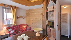 Ein gemütliches Zimmer mit Holzbalkendecke, einem rot gemusterten Sofa, einem Holztisch mit Tassen und einem Fernseher auf einer Holzkommode.