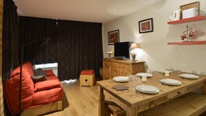 A cozy living room with a red sofa, a wooden dining table set for four, a television, and a cabinet with decorative items.