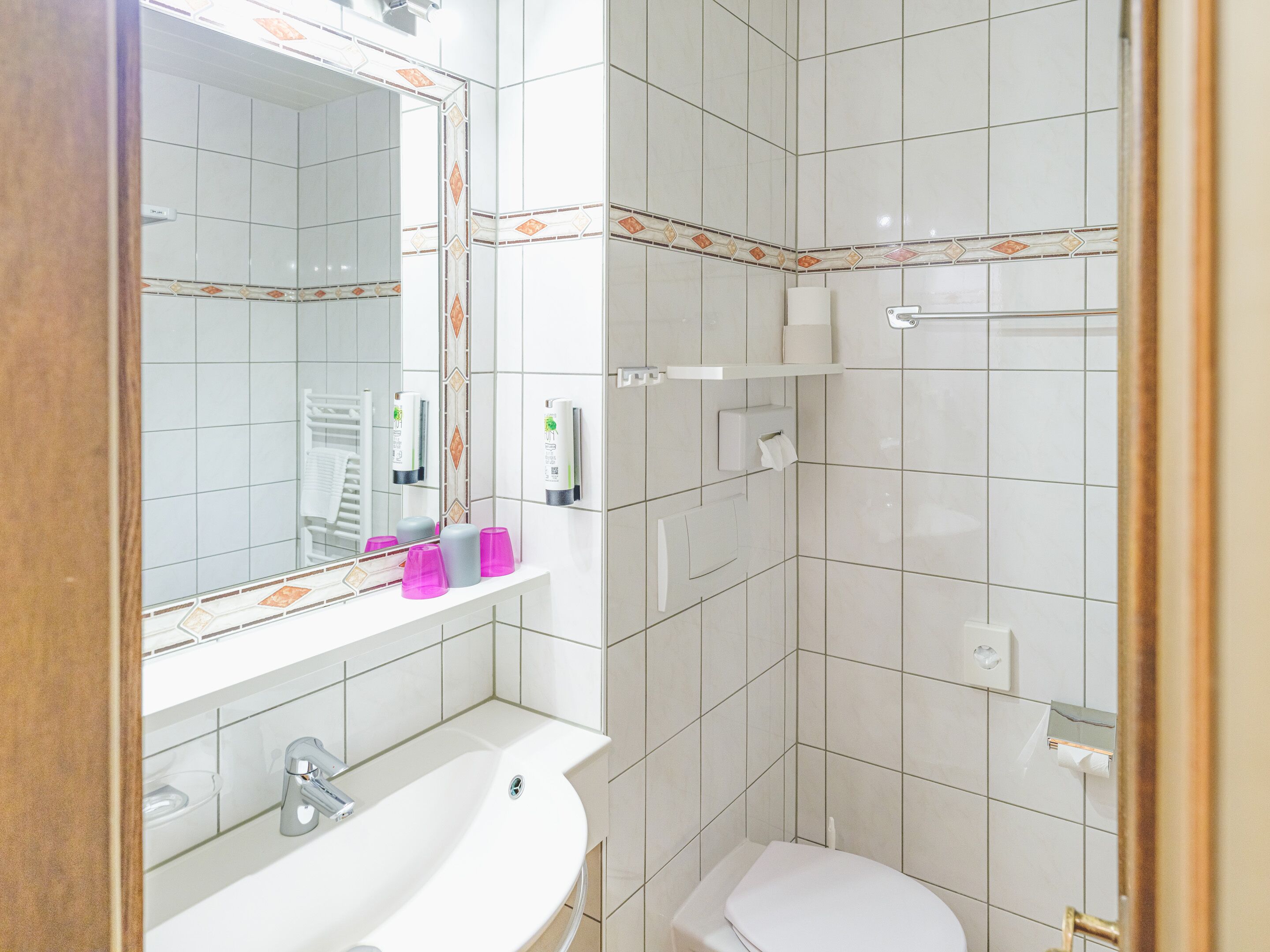 A bathroom with a white sink, mirror, and toilet. There are shelves with toiletries and a towel rack.