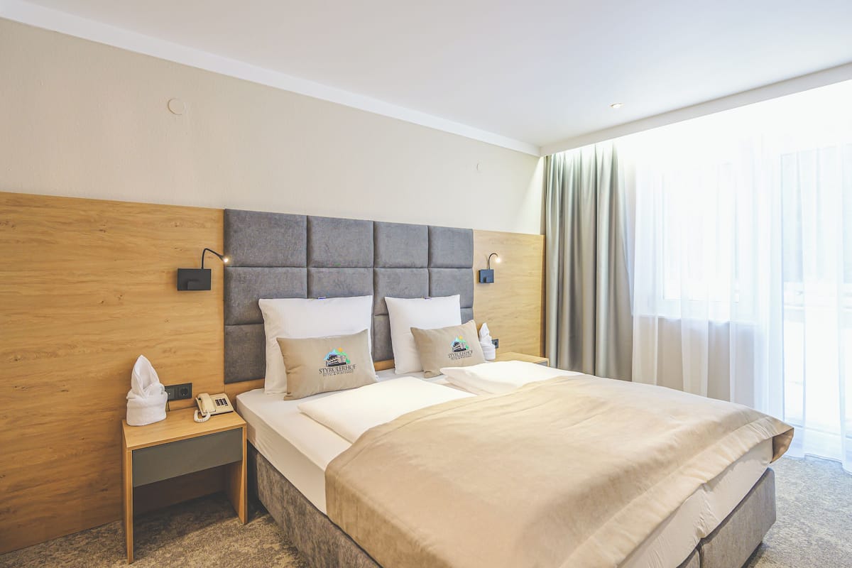 Hotellihuone, jossa on puinen sängynpääty, kaksi sänkyä, yöpöytä, jossa on puhelin ja paperiliinoja, sekä suuri ikkuna, jossa on verhot.
