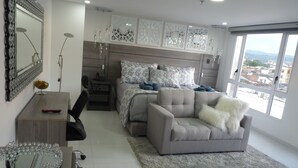 A modern bedroom with a large bed, a grey sofa, a desk, and a large window offering a city view.