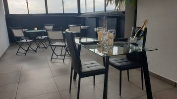 Une terrasse sur le toit avec une table en verre dressée pour le dîner, entourée de chaises noires, et une bouteille de champagne.