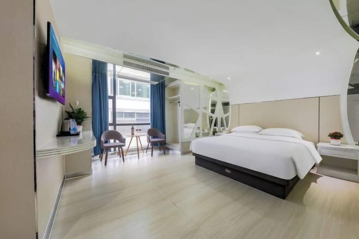 A modern hotel room with a large bed, a desk, and a chair. There is a window with curtains and a view of the outside.