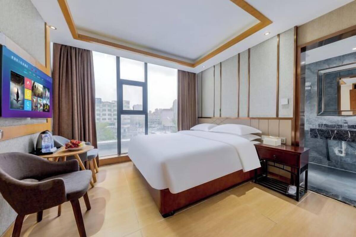 A hotel room with a bed, a desk, a chair, a TV, and a large window with a city view.