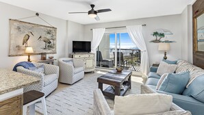 A spacious living room with a ceiling fan, a television, a dining area with a table and chairs, and a balcony with a view. - Gulf Views, Beach Chairs& Wagon, Steps to Pool& Beach, Spring Break Discount (Orange Beach)