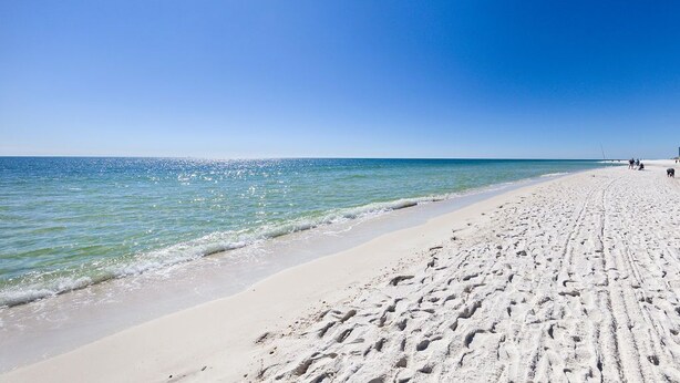 A wide sandy beach with footprints leading to the water, a clear blue sky, and a calm sea. - Gulf Views, Beach Chairs& Wagon, Steps to Pool& Beach, Spring Break Discount (Orange Beach)