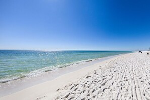 A wide sandy beach with footprints leading to the water, a clear blue sky, and a calm sea. - Gulf Views, Beach Chairs& Wagon, Steps to Pool& Beach, Spring Break Discount (Orange Beach)