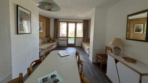 A dining area with a long table, chairs, and a lamp. There are windows with curtains, a sofa, and a bed in the background. - Ski-in/ski-out & foot of the slopes, 2nd floor, view ski slopes, balcony, tv, 35m², La Plagne (La Plagne)