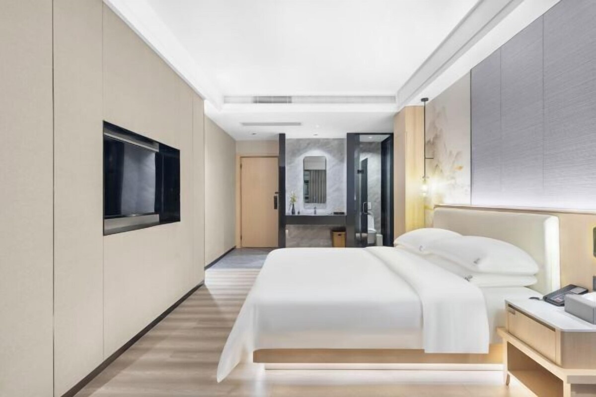 A modern hotel room with a large bed, a bedside table, a TV mounted on the wall, and a view into the bathroom.
