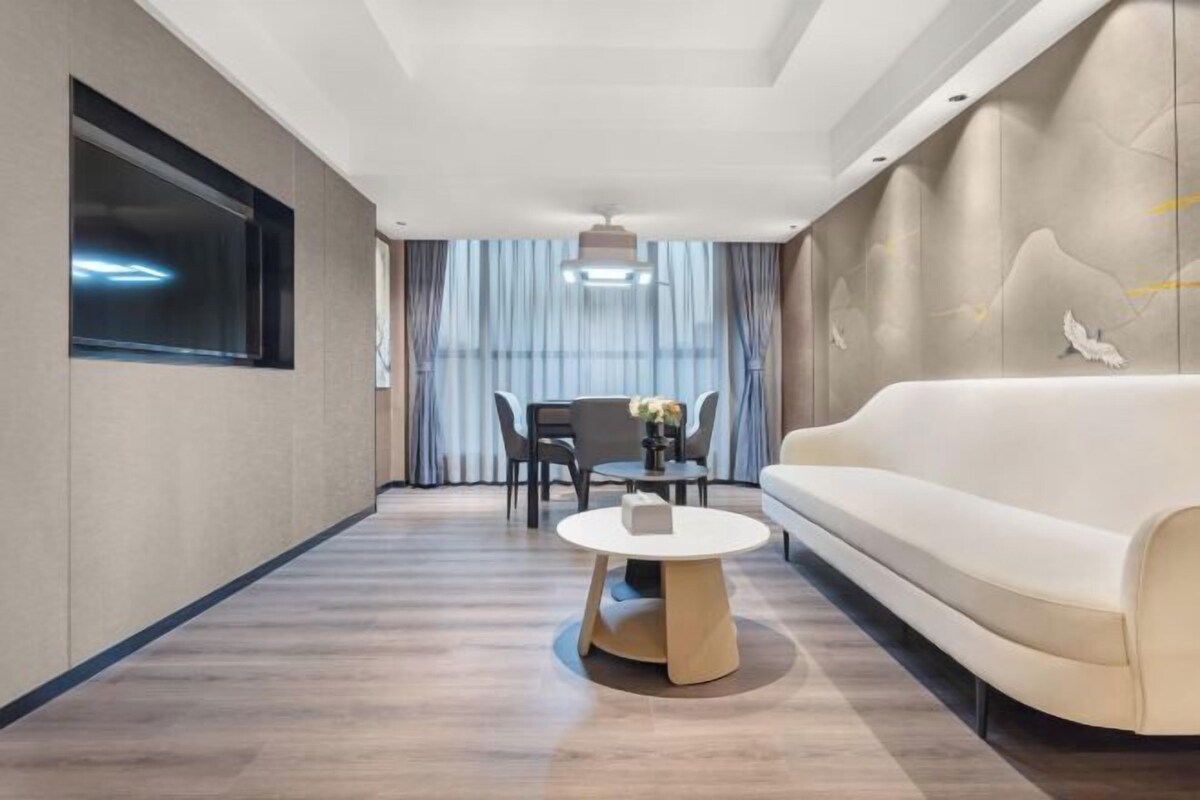 A modern hotel room with a sofa, a round coffee table, a dining table with chairs, and a large window with curtains.