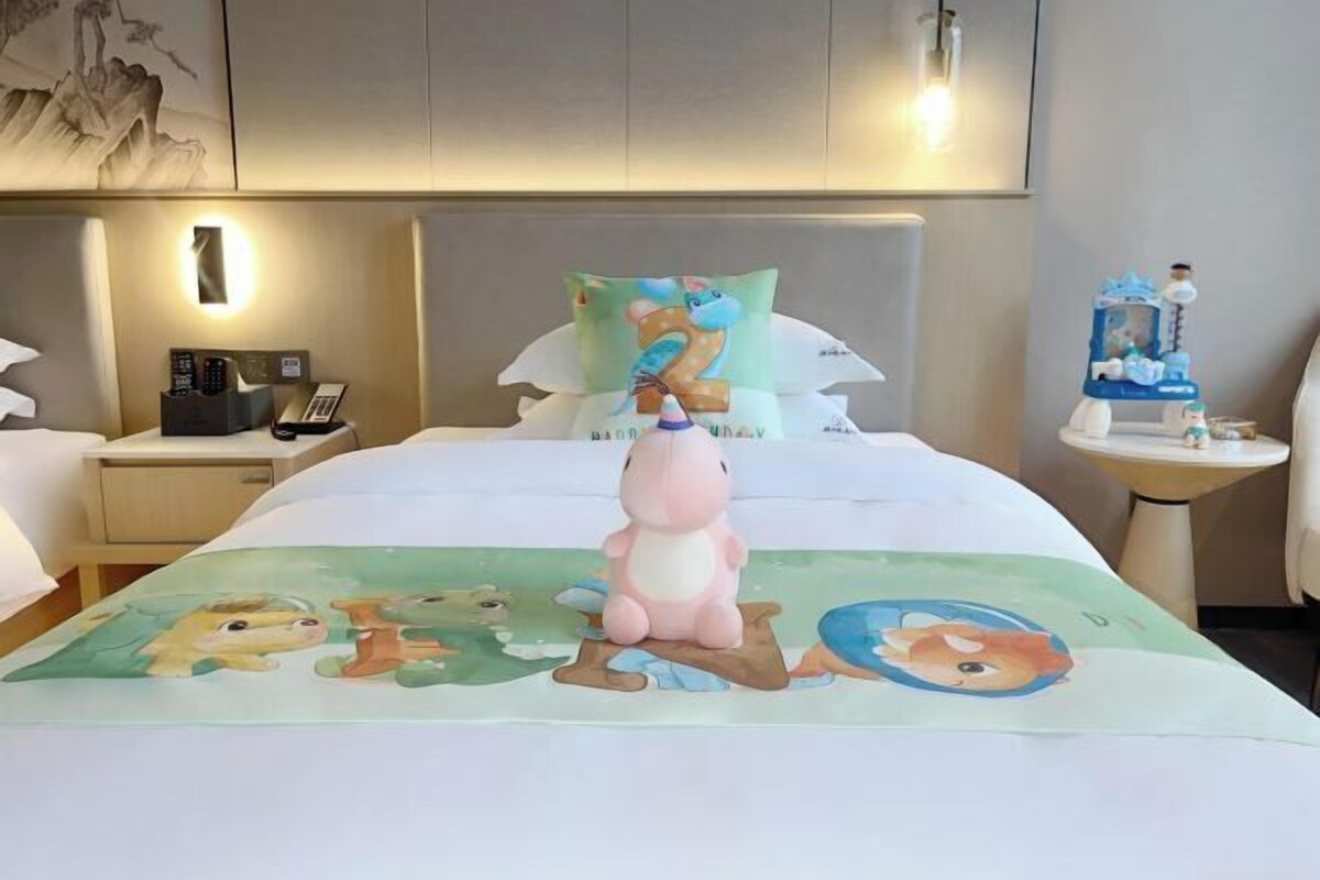 A hotel room with a bed featuring a cartoon animal-themed bedspread, a bedside table with a phone and a small decorative item, and a wall-mounted light fixture.