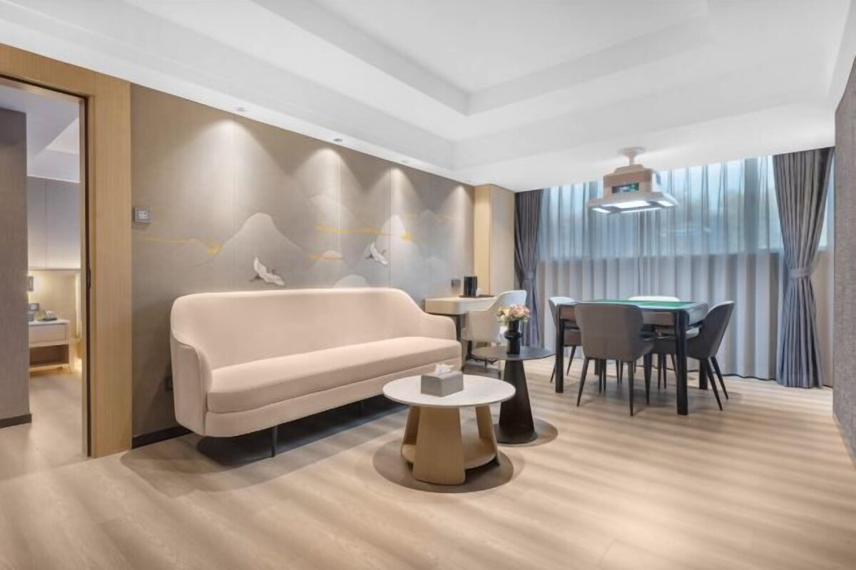 A modern hotel room with a sofa, a round coffee table, a dining table with chairs, and a large window with curtains.