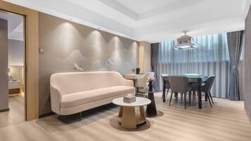 A modern hotel room with a sofa, a round coffee table, a dining table with chairs, and a large window with curtains.