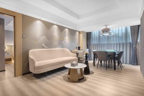 A modern hotel room with a sofa, a round coffee table, a dining table with chairs, and a large window with curtains.