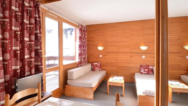 A cozy room with a wooden interior, a sofa bed, a dining table with chairs, and a window with curtains.