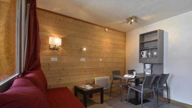 Una habitación acogedora con pared de madera, un sofá rojo, una mesita con una lámpara, y un comedor con sillas y una cafetera.