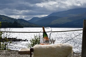 En flaska champagne, ett glas och en stol klädd i päls, placerade vid en sjö med berg i bakgrunden.
