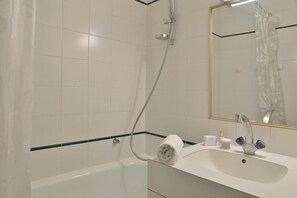 Ein modernes Badezimmer mit weißen Fliesen, einer Badewanne, einer Dusche inklusive Duschbrause, einem Waschbecken mit Spiegel und einem Handtuch am Badewannenrand.