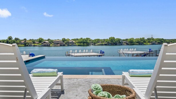 A poolside view with lounge chairs, a floating dock, and a view of a lake with buildings in the distance. - Waterhouse at Lake LBJ Residence II, 6 King Bedrooms|  Pool, Spa, Boat & PWC Lift, Slide  (Kingsland)
