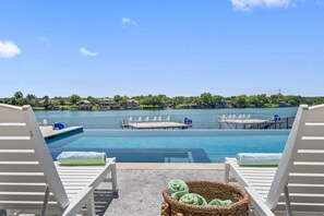 A poolside view with lounge chairs, a floating dock, and a view of a lake with buildings in the distance. - Waterhouse at Lake LBJ Residence II, 6 King Bedrooms|  Pool, Spa, Boat & PWC Lift, Slide  (Kingsland)