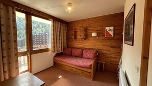 A cozy room with a wooden sofa, a small table, a window with curtains, and a wall-mounted shelf. - 100m from ski slopes, 8th floor, view ski slopes, balcony, tv, ski locker, 28m², La Plagne (La Plagne)