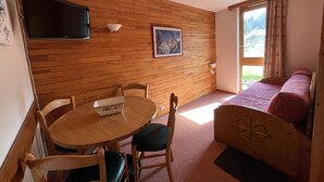 A room with a wooden wall, a round table with chairs, a bed with a headboard, a TV mounted on the wall, and a window with curtains. - 100m from ski slopes, 8th floor, view ski slopes, balcony, tv, ski locker, 28m², La Plagne (La Plagne)
