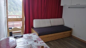 A small room with a wooden bed, a table with a tablecloth, a red curtain, and a view of a mountain through a window. - 100m from ski lifts, 7th floor, balcony, tv, ski locker, 15m², La Plagne (La Plagne)
