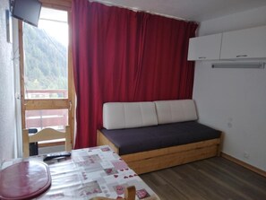 A small room with a wooden bed, a table with a tablecloth, a red curtain, and a view of a mountain through a window. - 100m from resort center, 7th floor, balcony, tv, ski locker, 15m², La Plagne (La Plagne)