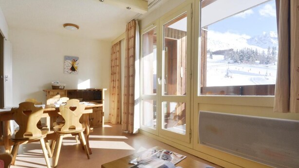 A room with a wooden dining table and chairs, a sofa, and a large window with a view of a snowy landscape.