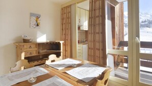 A dining area with a wooden table set for four, a wooden cabinet with drawers, a painting on the wall, and a view of a snowy landscape through a glass door.