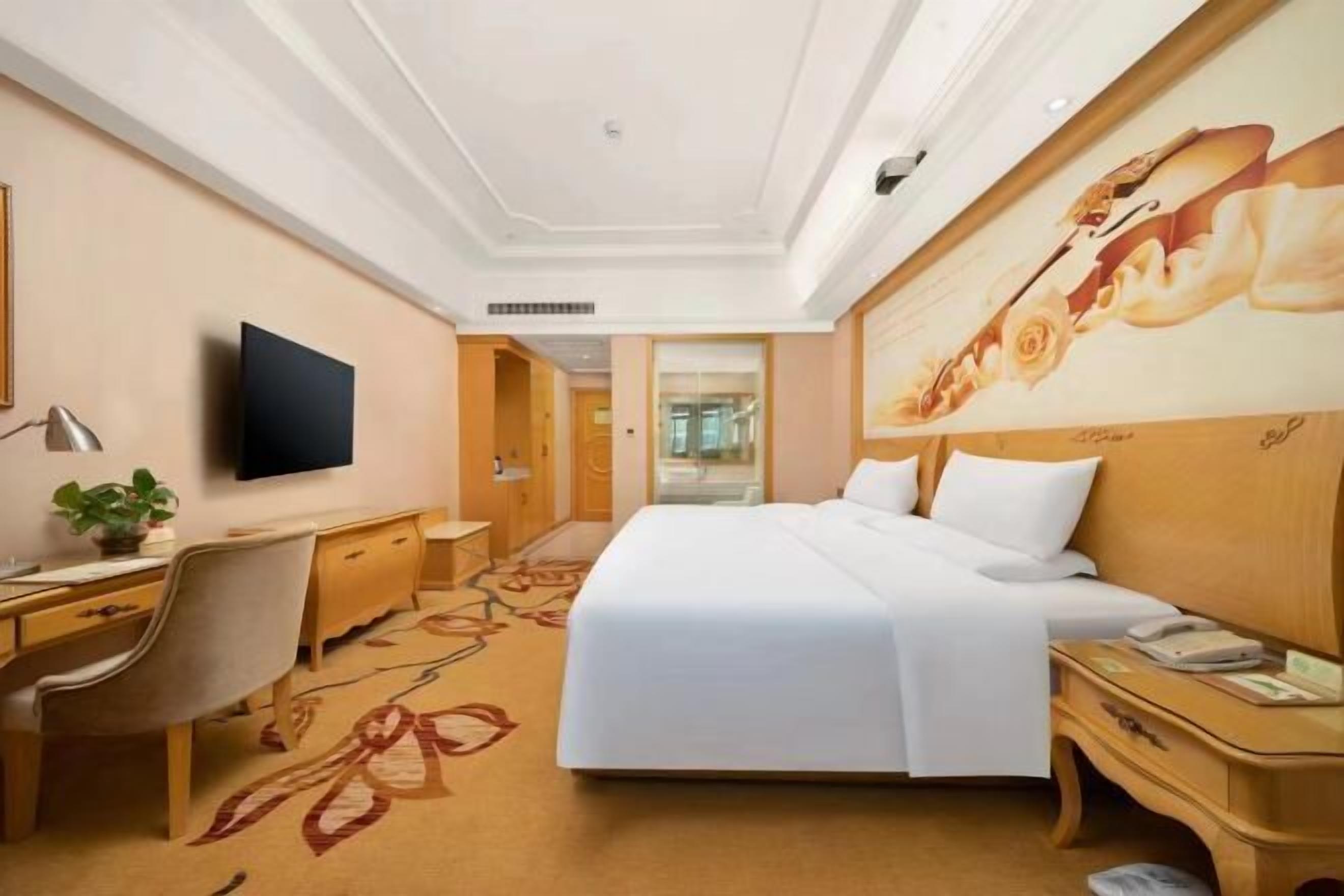 A hotel room with a large bed, a desk with a chair, a television, and a painting on the wall.
