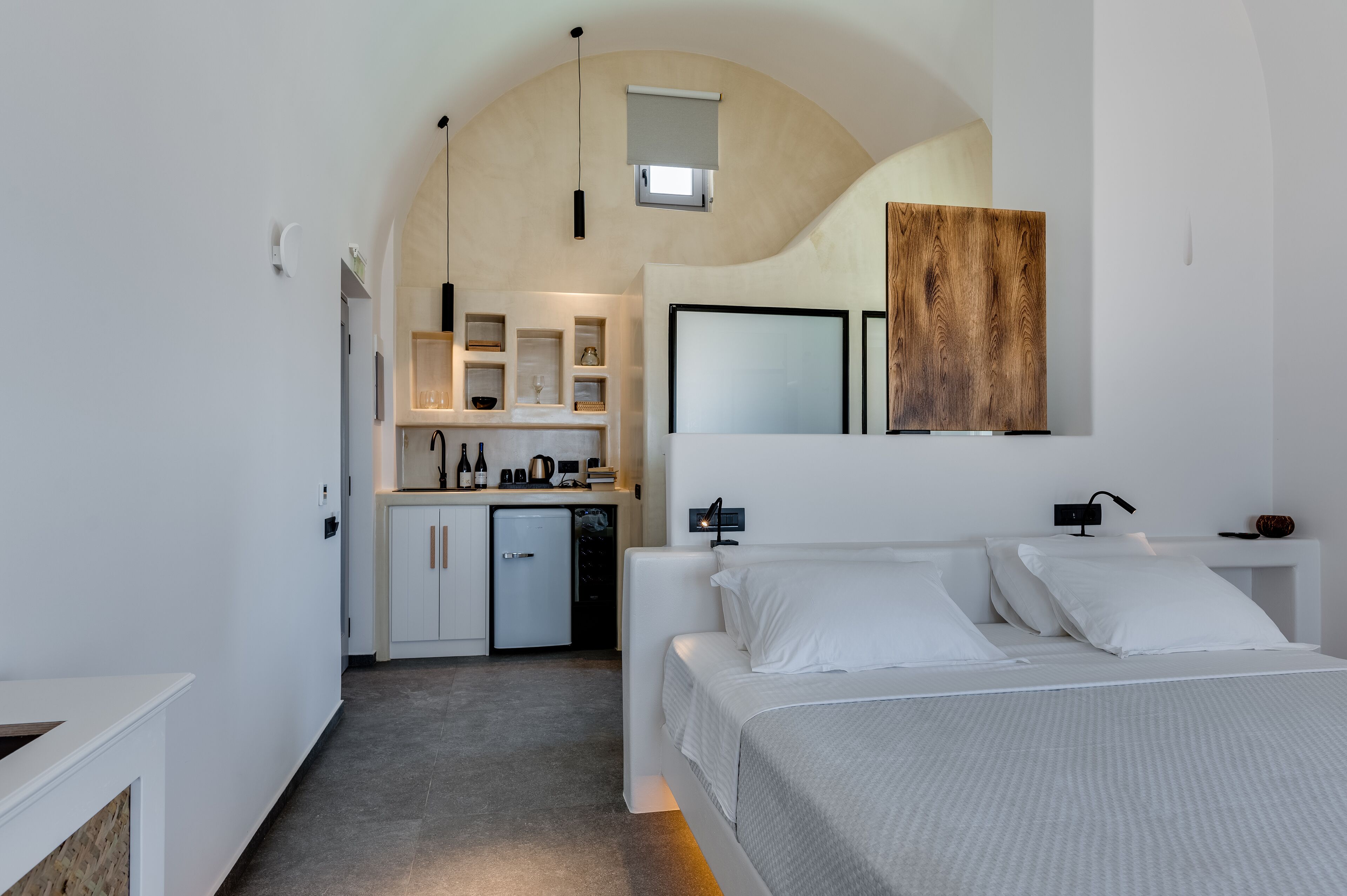 A modern hotel room with a minimalist design, featuring a large bed, a small kitchenette, and a unique arched ceiling.